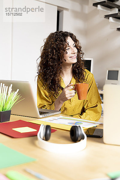 Lächelnde Frau mit Tasse Kaffee sitzt am Schreibtisch im Büro