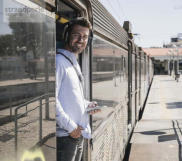 Lächelnder junger Mann mit Kopfhörern und Smartphone steht in der Zugtür