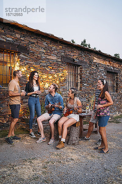 Gruppe von Freunden spielt in der Abenddämmerung Ukulele im Freien an einem Steinhaus