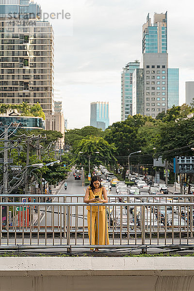 Modisch gekleidete Frau in Gelb steht auf einer Brücke und schaut auf Handy  Bangkok  Thailand