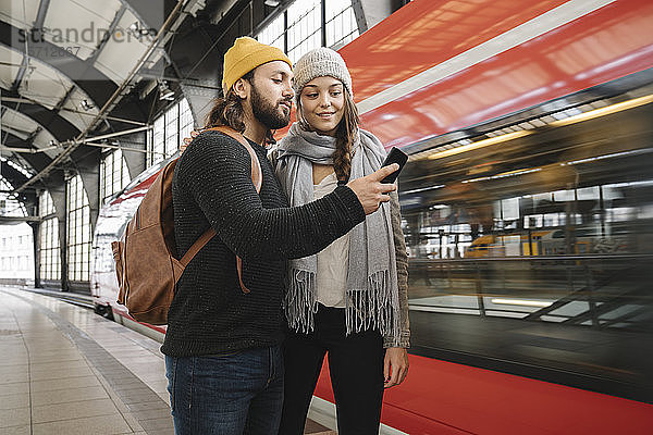 Junges Paar benutzt ein Smartphone am Bahnsteig  als der Zug einfährt  Berlin  Deutschland