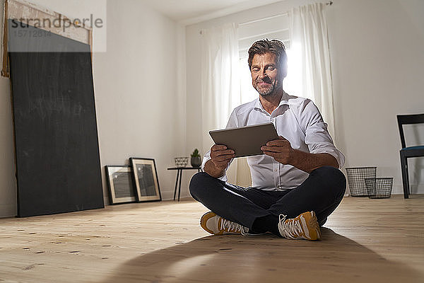Porträt eines lächelnden  reifen Mannes  der zu Hause auf dem Boden sitzt und ein digitales Tablet benutzt