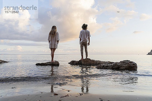 Rückenansicht eines jungen Paares  das auf Felsen vor dem Meer steht und ein Flugzeug beobachtet  Ibiza  Balearen  Spanien