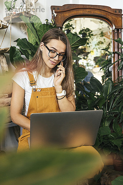 Junge Frau mit Laptop am Telefon in einem kleinen Gartenbaubetrieb