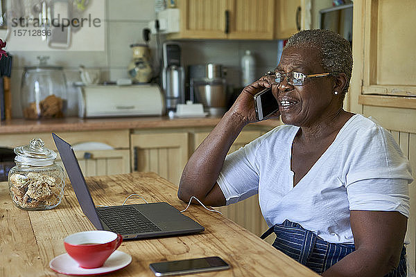 Ältere Frau sitzt am Küchentisch mit Laptop und Smartphone