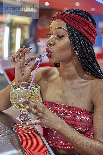 Junge Frau mit geflochtener Frisur sitzt an der Bar eines Restaurants und trinkt einen Cocktail mit einem Strohhalm