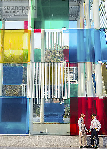 Elegantes schwules Paar hält sich an einem Gebäude mit farbenfroher Fassade an den Händen