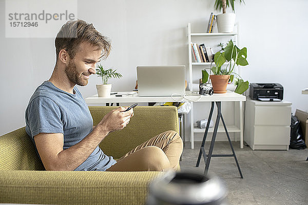 Lächelnder Mann sitzt im Büro auf der Couch und benutzt ein Smartphone