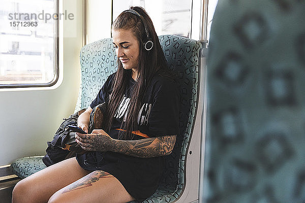 Tätowierte junge Frau sitzt mit Smartphone und Kopfhörern in der Pendlerschlange  Berlin  Deutschland