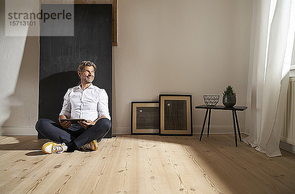 Entspannter  reifer Mann mit digitalem Tablett  der zu Hause auf dem Boden sitzt und aus der Ferne schaut
