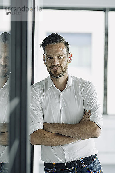 Porträt eines selbstbewussten  reifen Geschäftsmannes  der sich im Büro an eine Glaswand lehnt