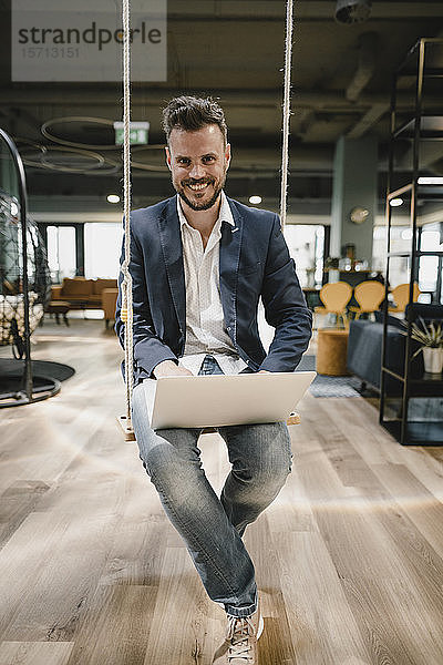 Geschäftsmann mit Laptop im Coworking Space  auf der Schaukel sitzend