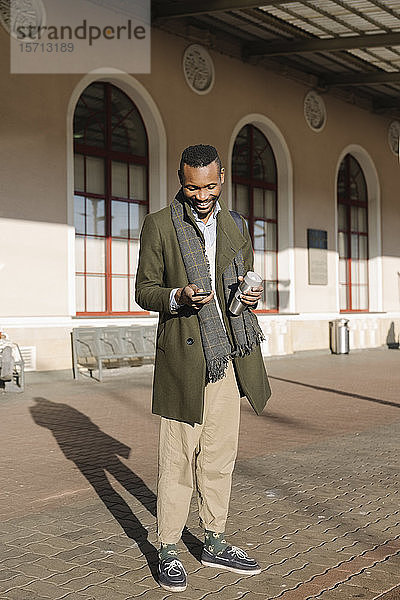 Stylischer Mann mit wiederverwendbarem Becher  der während des Wartens auf den Zug ein Smartphone benutzt