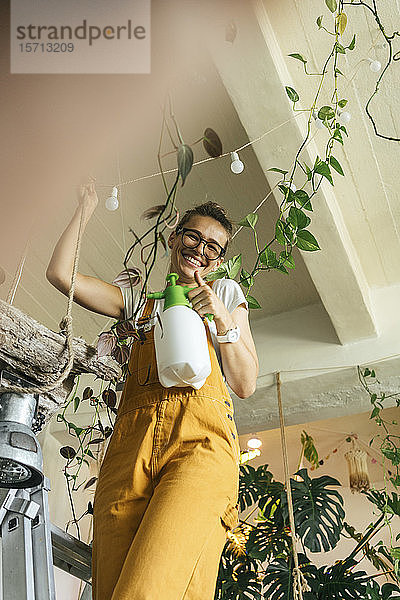 Glückliche junge Frau steht auf einer Leiter und kümmert sich in einem kleinen Laden um Pflanzen
