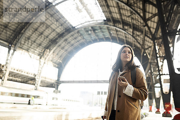 Junge Frau mit Handy am Bahnhof  die sich umsieht
