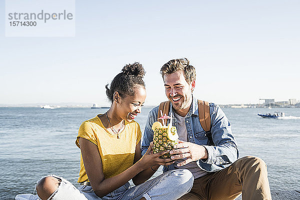Glückliches junges Paar sitzt am Wasser und trinkt ein Getränk in einer Ananas  Lissabon  Portugal