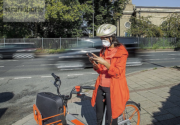 Junge Frau mit Helm und Gesichtsmaske  auf Fahrrad sitzend  mit Smartphone