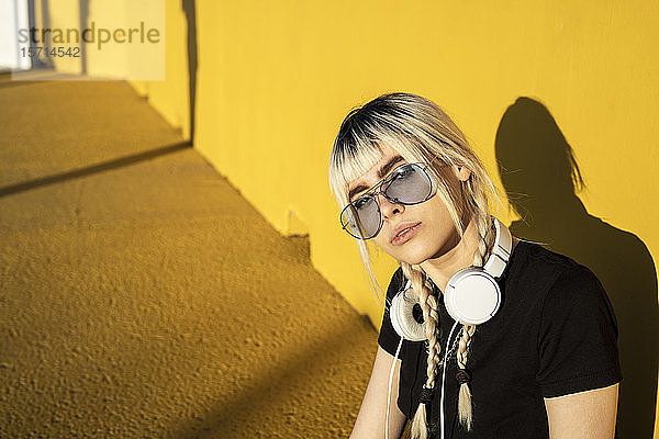 Porträt einer jungen Frau mit Kopfhörer und farbiger Sonnenbrille vor gelber Wand