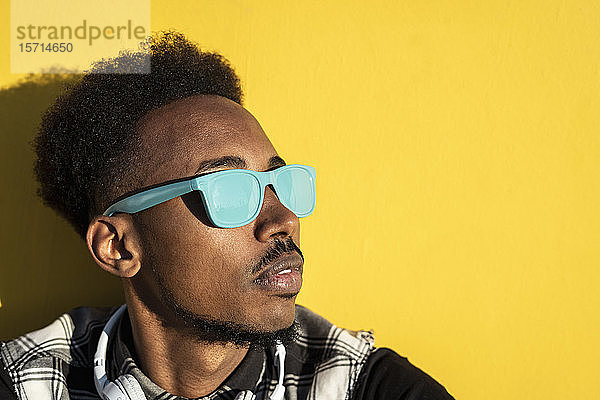Porträt eines jungen Mannes mit hellblauer Sonnenbrille vor gelber Wand