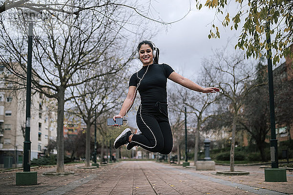 Porträt einer jungen Frau in schwarzer Sportkleidung  die in die Luft springt  während sie mit Kopfhörern Musik hört