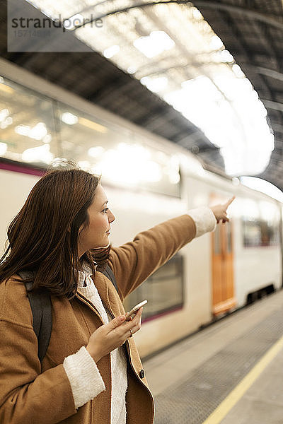Junge Frau mit Handy auf dem Bahnhof  die mit dem Finger zeigt