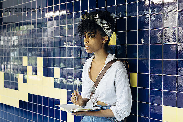 Porträt einer jungen Frau mit Notizbuch  das an einer gekachelten Wand lehnt und in die Ferne schaut