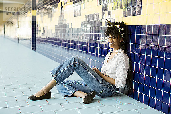 Porträt einer glücklichen jungen Frau  die mit einem Smartphone auf dem Fliesenboden sitzt