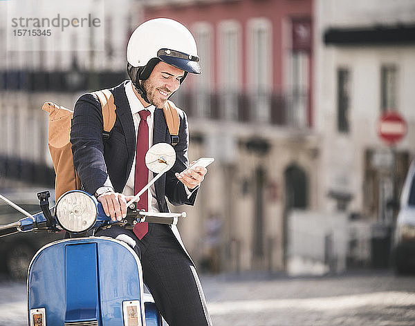 Lächelnder junger Geschäftsmann auf Motorroller mit Handy in der Stadt  Lissabon  Portugal