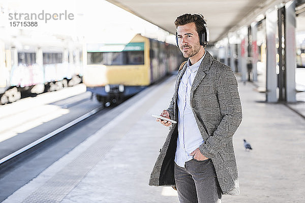 Junger Geschäftsmann mit Handy und Kopfhörern am Bahnhof