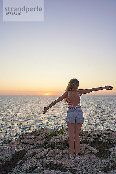 Junge Frau steht auf einer Klippe  schaut auf den Sonnenuntergang  Rückansicht