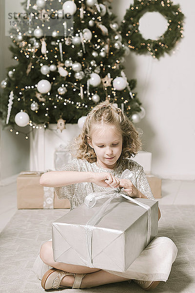 Porträt eines blonden kleinen Mädchens  das vor dem Weihnachtsbaum sitzt und das Weihnachtsgeschenk öffnet
