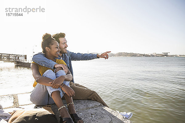 Glückliches junges Paar sitzt auf dem Pier am Wasser und schaut auf die Aussicht  Lissabon  Portugal