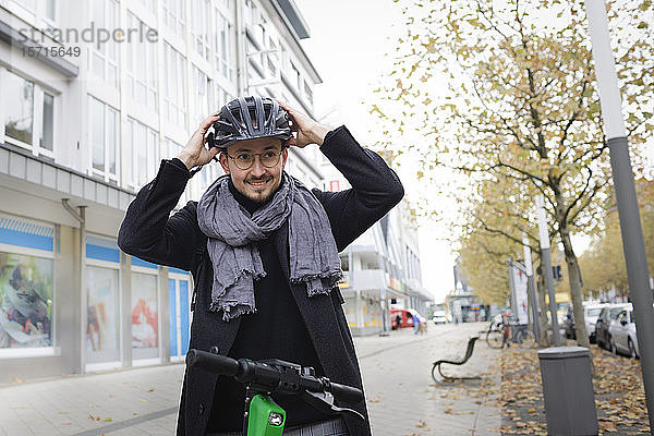 Porträt eines lächelnden jungen Mannes  der mit einem Elektroroller auf dem Bürgersteig steht und einen Fahrradhelm aufsetzt