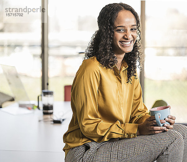 Porträt einer glücklichen jungen Geschäftsfrau  die im Büro am Schreibtisch sitzt und eine Kaffeepause macht