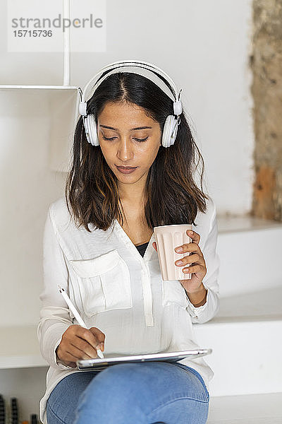 Porträt eines jungen Architekten mit Kopfhörer und Kaffeetasse  der auf Stufen in einem Atelier sitzt und an einem Tablett arbeitet