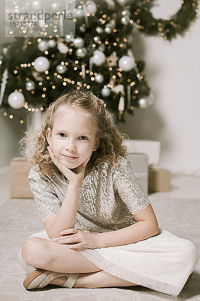 Porträt eines blonden kleinen Mädchens vor dem Weihnachtsbaum sitzend