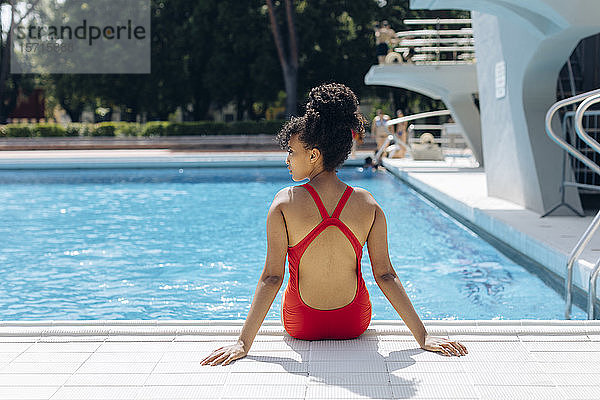 Rückenansicht einer jungen Frau im roten Badeanzug am Poolufer sitzend