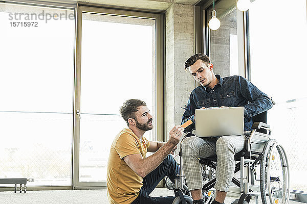 Junger Geschäftsmann im Rollstuhl und Kollege mit Laptop im Büro