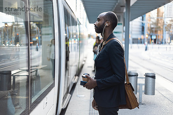 Junger Geschäftsmann mit Kaffee zum Mitnehmen steht an Straßenbahnhaltestelle und hört mit Kopfhörern und Smartphone Musik