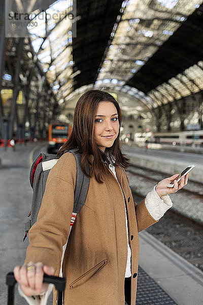 Porträt einer lächelnden jungen Frau mit Handy in der Hand am Bahnhof