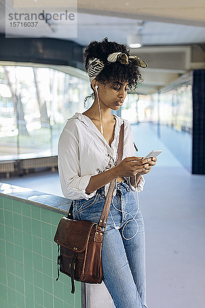 Porträt einer jungen Frau mit Ohrstöpseln  die in einer Passage auf ein Handy schaut
