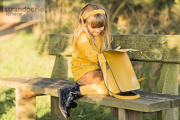Kleines blondes  gelb gekleidetes Mädchen auf Gartenbank sitzend mit Ledertasche