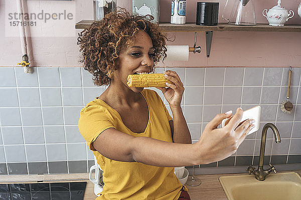 Junge Frau  die einen Maiskolben isst und einen Selfie in der Küche nimmt