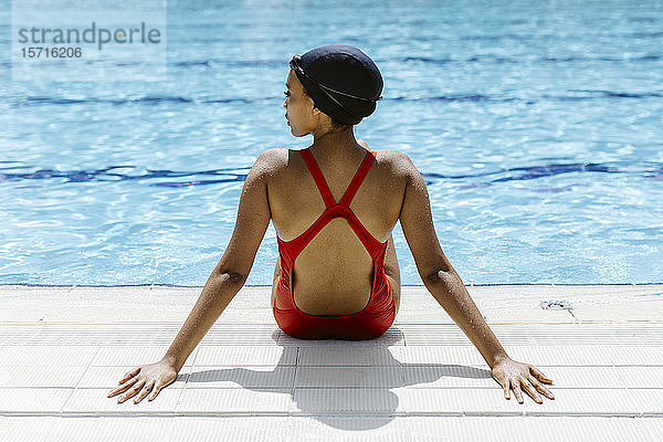 Rückenansicht einer jungen Frau in rotem Badeanzug und Badekappe  die sich nach dem Schwimmen am Pool entspannt