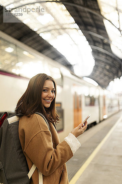Porträt einer lächelnden jungen Frau mit Handy auf dem Bahnhof