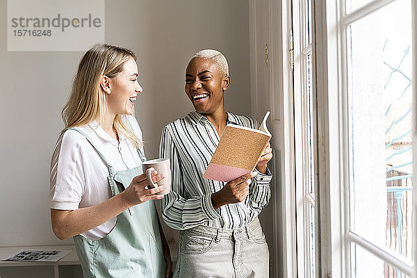 Zwei glückliche Frauen mit Notebook und Tasse Kaffee am Fenster