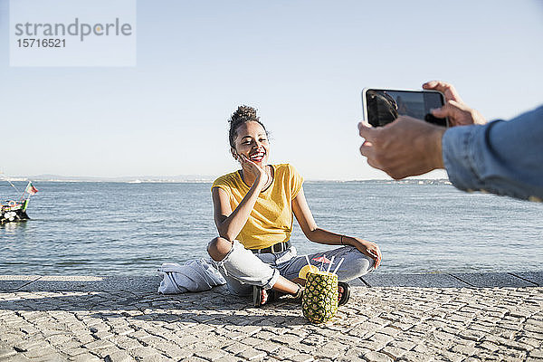 Junger Mann beim Fotografieren seiner Freundin  die am Pier am Wasser sitzt  Lissabon  Portugal
