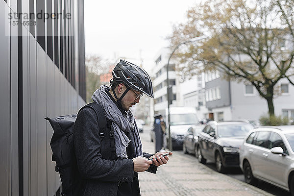 Junger Mann mit Fahrradhelm und Rucksack steht auf dem Bürgersteig und schaut auf sein Handy