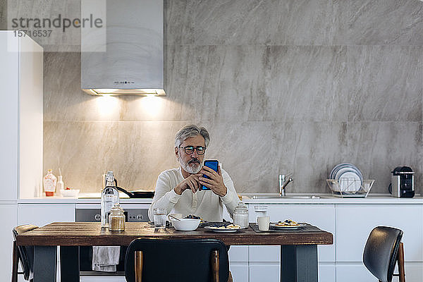 Reifer Mann sitzt zu Hause am Tisch in der Küche und benutzt sein Handy