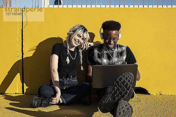 Porträt eines lachenden jungen Paares  das mit Kopfhörer und Laptop vor der gelben Wand sitzt